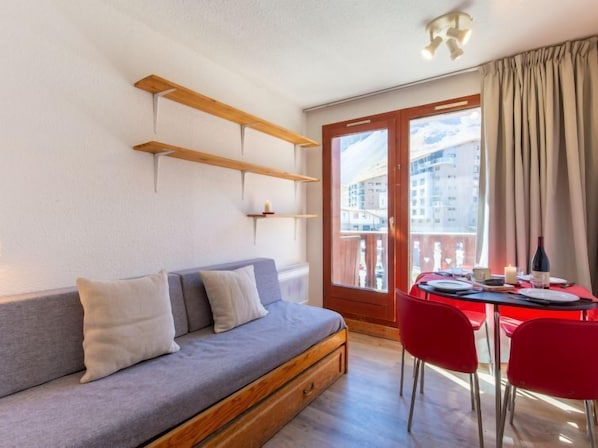A modern living room with a grey sofa, wooden shelves, a dining table with red chairs, and a large window with a view of buildings. - Chalet Club (Val Claret) by Interhome (Tignes)