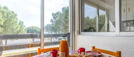 A dining table set with red tableware, a bottle of orange juice, and pastries, with a view of trees and a road through large windows.
