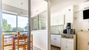 Cocina moderna con armarios blancos, microondas y cafetera. Cuenta con un comedor con mesa y sillas, y un ventanal corredero que da a un balcón con vistas a árboles.