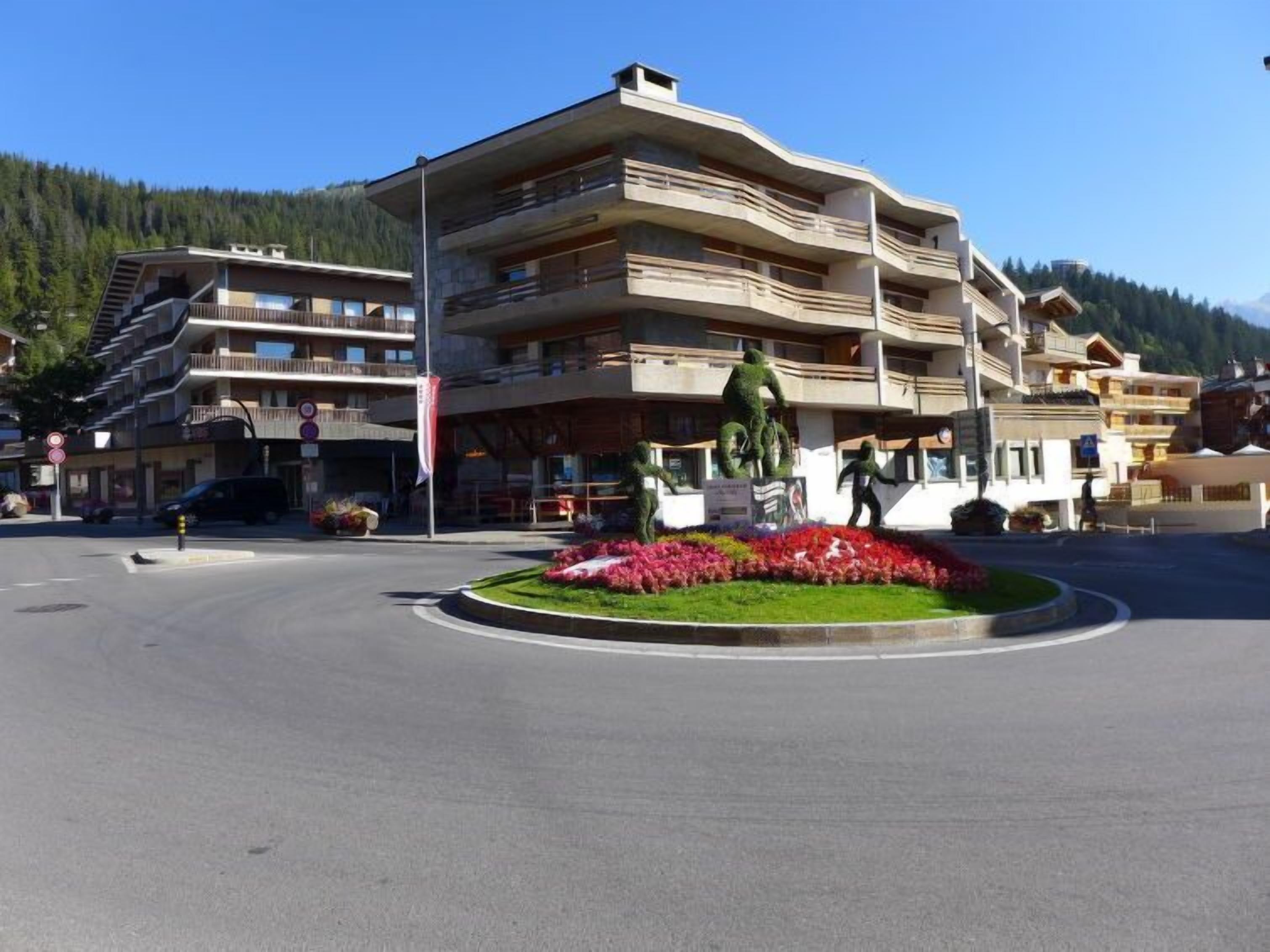 A multi-story hotel with balconies, a central flower bed, and a mountainous backdrop.