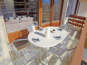 Une table ronde entourée de quatre chaises, un pichet blanc, deux tasses et un petit récipient, le tout sur un balcon doté de rampes en verre et de mobilier en bois.