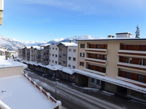 A row of multi-story buildings with balconies, a snow-covered street, and a mountain backdrop.