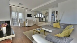 Ein modernes Wohnzimmer mit einer Couch, einem Couchtisch und einem Fernseher. Im Hintergrund ist eine Küche mit einem Kühlschrank sowie ein Essbereich mit Stühlen zu sehen.