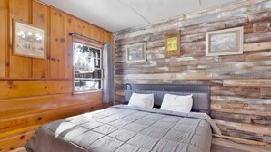 A bedroom with a wooden paneled wall, a bed with a grey quilt, and framed pictures on the wall. - Juniper #105 (Big Bear Lake)