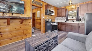 A cabin interior with wooden walls, a kitchen with a stove, microwave, and refrigerator, a gray sofa, and a mounted television displaying a scenic landscape. - Juniper #105 (Big Bear Lake)