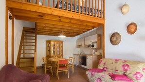 Un salon chaleureux avec un escalier en bois, une table à manger avec des chaises, un canapé recouvert d’un tissu à motifs floraux et un coin cuisine équipé de placards et d’un four à micro-ondes.