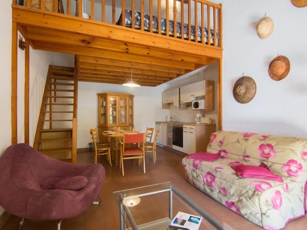 A cozy living area with a wooden staircase, a dining table with chairs, a sofa with floral upholstery, and a kitchen area with cabinets and a microwave.