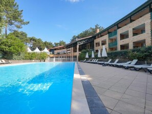 Ein Schwimmbecken mit Liegestühlen und Sonnenschirmen, umgeben von einem Gebäude mit mehreren Balkonen und blauem Himmel.