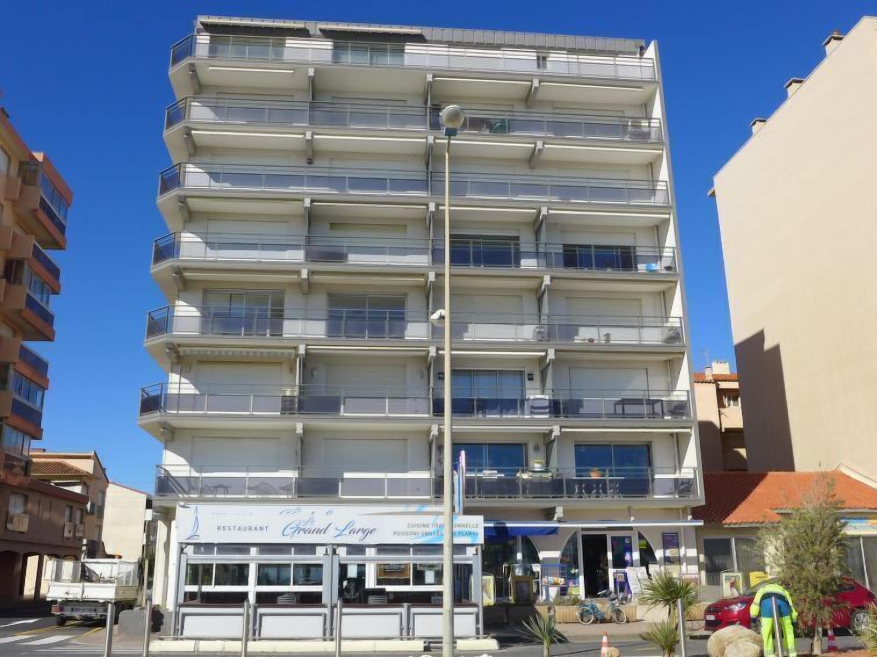 A multi-story hotel with balconies on each floor, a restaurant named "Grand Large" at the ground level, and a parking area with a few cars and a person in a high-visibility vest.