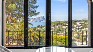 Ein Zimmer mit einem großen Bogenfenster, das Blick auf eine Küstenlandschaft bietet, darunter Bäume, ein Berg und ein Gewässer.