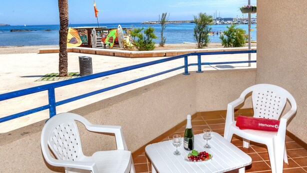 A balcony with two white plastic chairs, a small table, and a bottle of wine. The balcony overlooks a beach with a clear view of the sea and boats. - Maritim II by Interhome (Llançà)