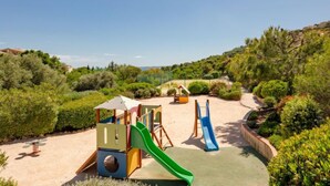 A playground with slides and climbing structures surrounded by greenery and hills.