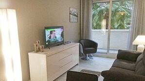 Ein Wohnzimmer mit Fernseher, einer Couch, einem Sessel und einem Fenster mit Blick auf Palmen.
