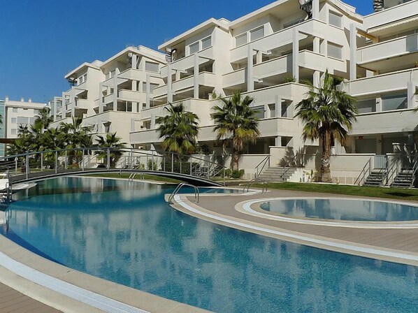 A modern hotel with a central swimming pool, multiple balconies, and palm trees.