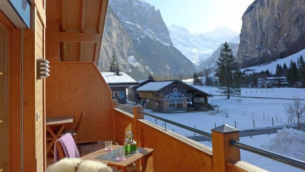 A wooden balcony with a table and chairs, overlooking a snowy mountain landscape with houses. - Staubbach by Interhome (Lauterbrunnen)