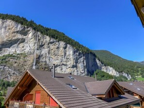 A chalet with a sloped roof, wooden exterior, and a red door, set against a backdrop of a steep cliff and forested mountain.