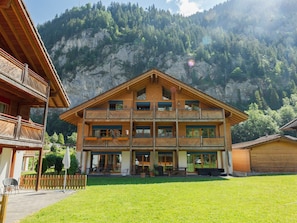 A multi-story wooden building with balconies, a covered outdoor seating area, and a mountain backdrop.