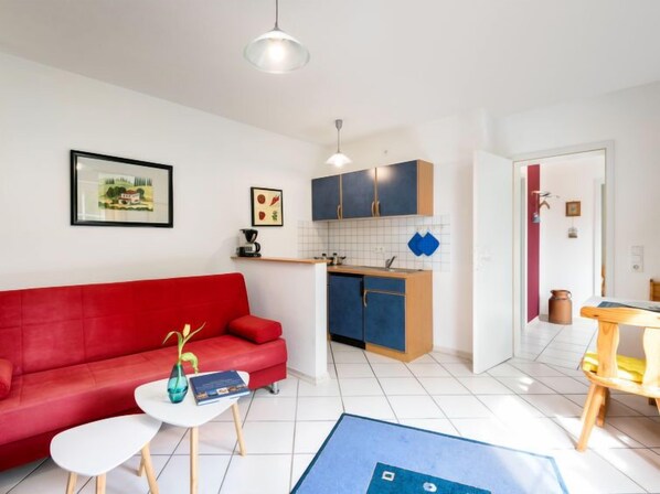 A modern living room with a red sofa, a small white table, and a blue rug. There is a kitchenette with blue cabinets and a white countertop.