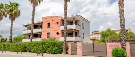 A multi-story residential building with balconies, surrounded by palm trees and a gated entrance.