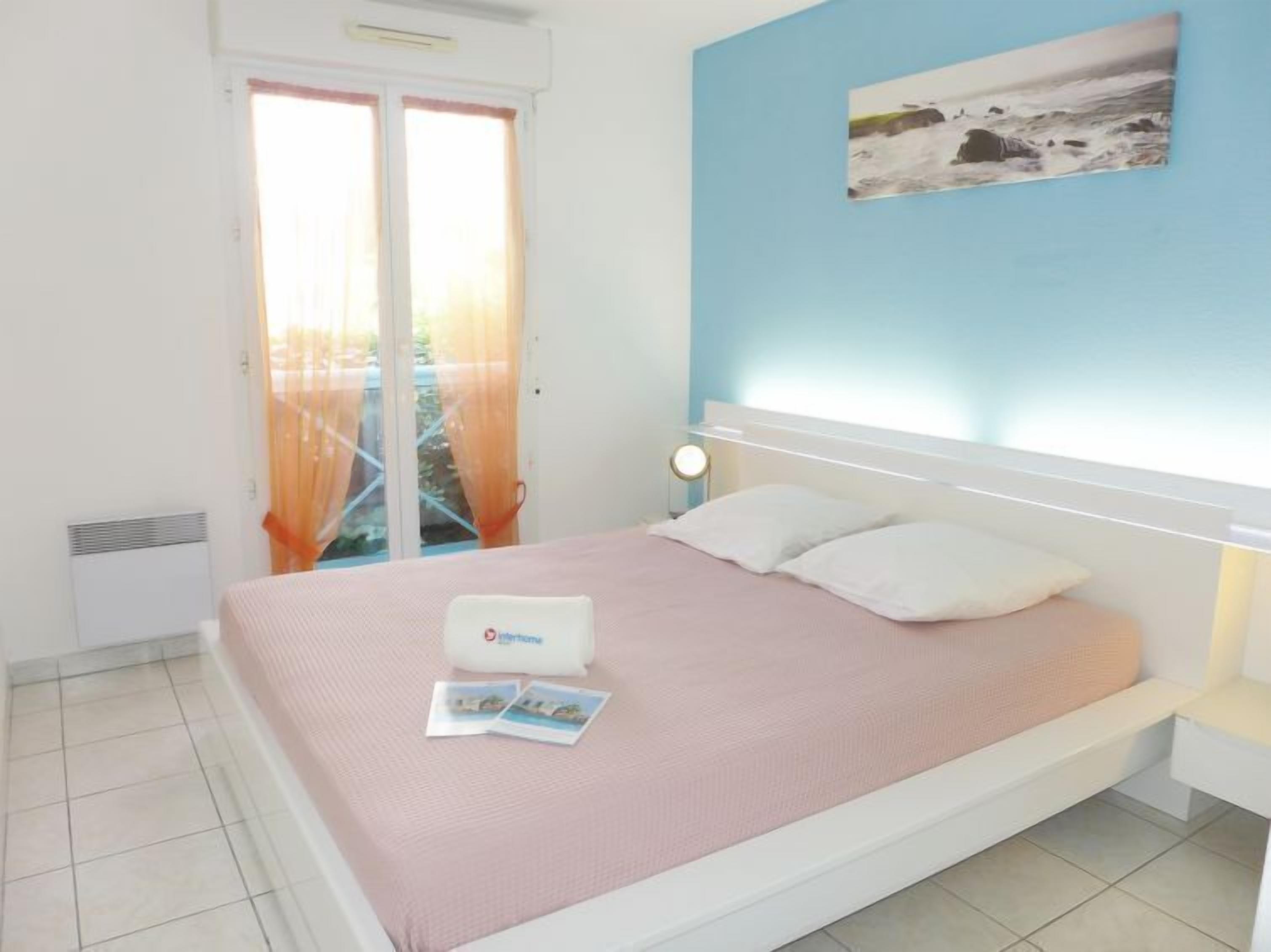 A bedroom with a bed, a nightstand, a wall-mounted lamp, a window with curtains, and a painting.