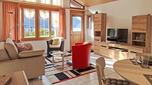 A modern living room with a sofa, armchair, and red chair, a TV, wooden cabinets, and large windows with curtains.