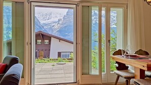 A room with a wooden floor, a dining table with chairs, a sofa, and a sliding glass door leading to a mountain view.