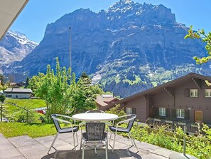 A patio with a table and chairs, a mountain backdrop, and a building with a sloped roof.