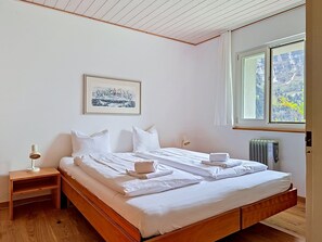 Une chambre à coucher avec un lit en bois, du linge de lit blanc, une table de chevet avec une lampe, un radiateur et une fenêtre donnant sur des arbres.