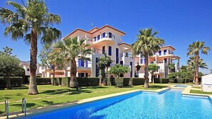 A swimming pool with a clear blue water surface, surrounded by a well-maintained lawn and palm trees. In the background, there are multi-story buildings with balconies and a clear blue sky. - Alma del Sol by Interhome (Dénia)