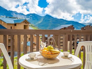 A table with a fruit basket and two cups on a balcony overlooking mountains.