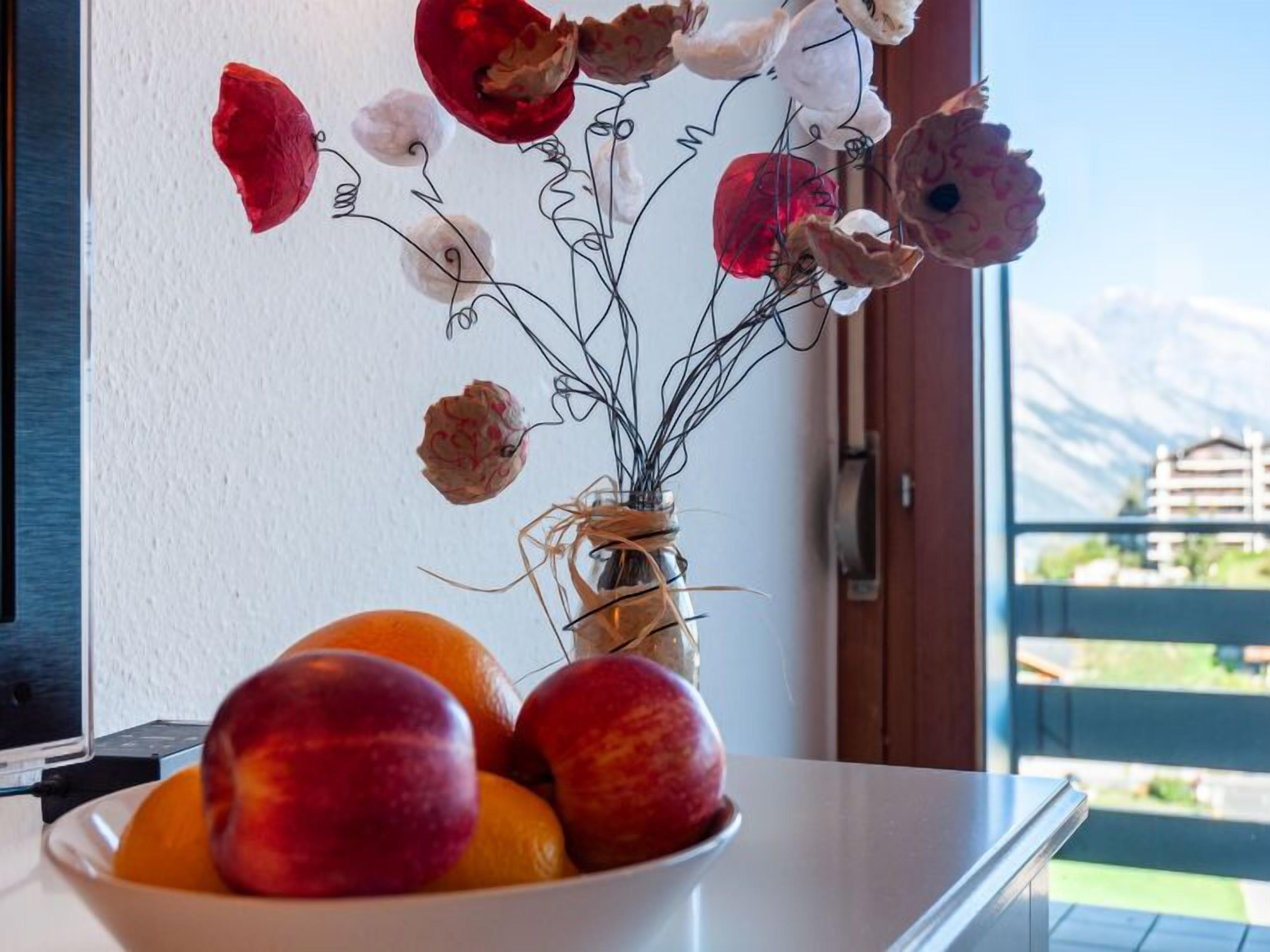 A bowl of fruit and a vase with dried flowers on a table by a window with a view of mountains.