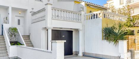Un edificio de dos pisos con fachada blanca, un balcón con barandilla y una zona de entrada con tejas.