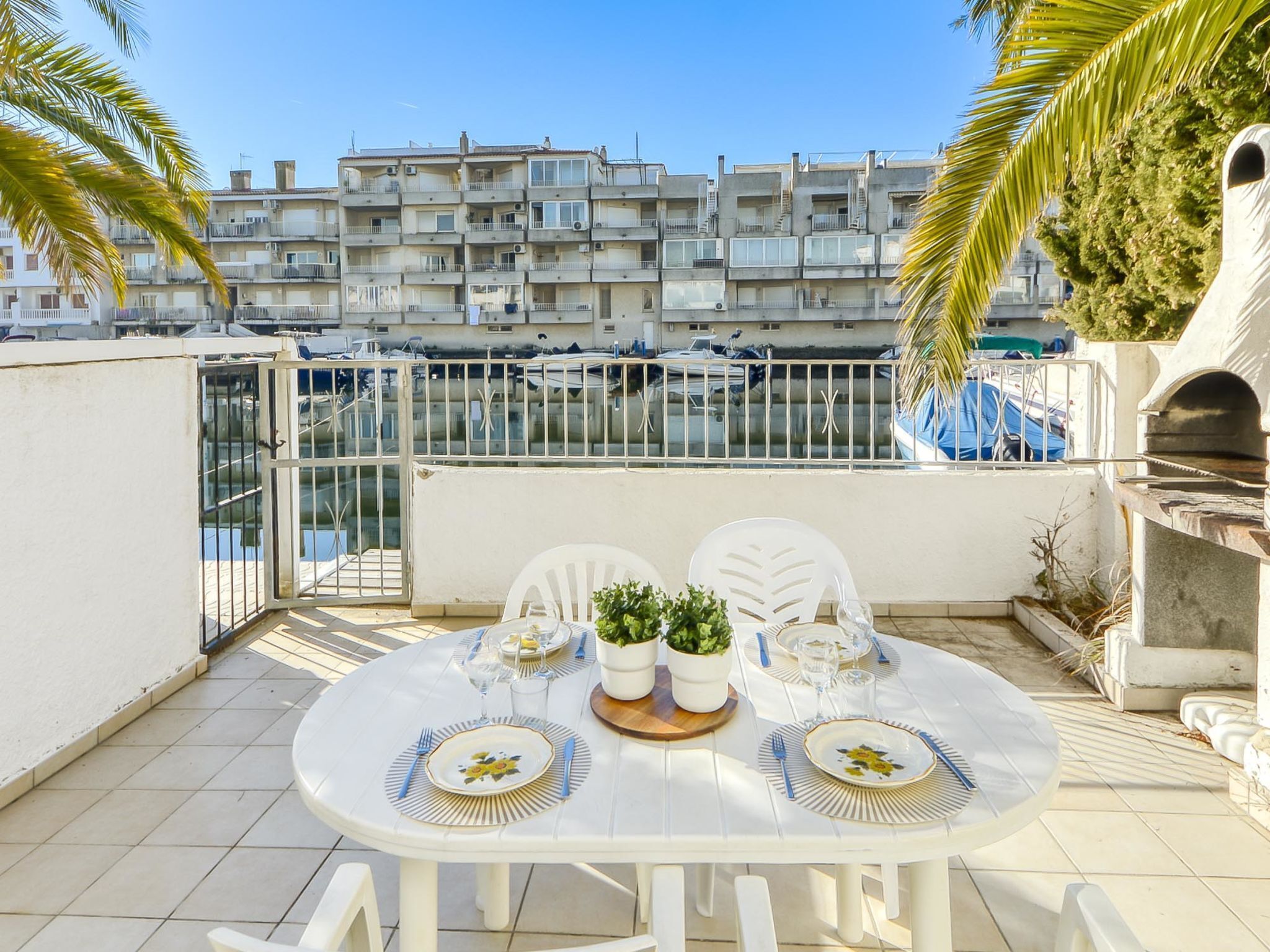 A white round table with chairs, a small potted plant, and a grill on a balcony with a view of a marina and apartment buildings.