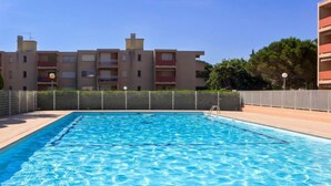 Une piscine rectangulaire à l’eau d’un bleu limpide, entourée d’une terrasse carrelée. Au fond, on aperçoit des immeubles à plusieurs étages, dotés de balcons, et une clôture.