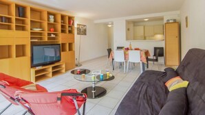 A living room with a sofa, a TV, a red chair, a glass table, and a dining area with a table and chairs. - Le Magellan by Interhome (Saint-Raphaël)