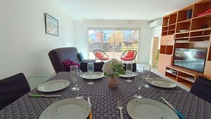 A dining area with a table set for four, chairs, a television, and a window with a view of trees. - Le Magellan by Interhome (Saint-Raphaël)