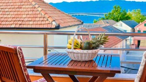 Un balcon avec un panier en osier rempli de fruits, une chaise en bois et une vue sur des maisons et des montagnes.
