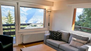 Una sala de estar moderna con un sofá gris, una mesa de centro de madera y grandes ventanales con vistas a un paisaje montañoso.