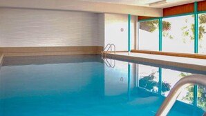 Una piscina interior con agua azul cristalina, una escalera metálica y ventanas amplias con vistas a la vegetación exterior.
