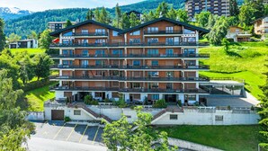 Un hotel de varios pisos con balcones, una zona de estacionamiento y montañas al fondo.