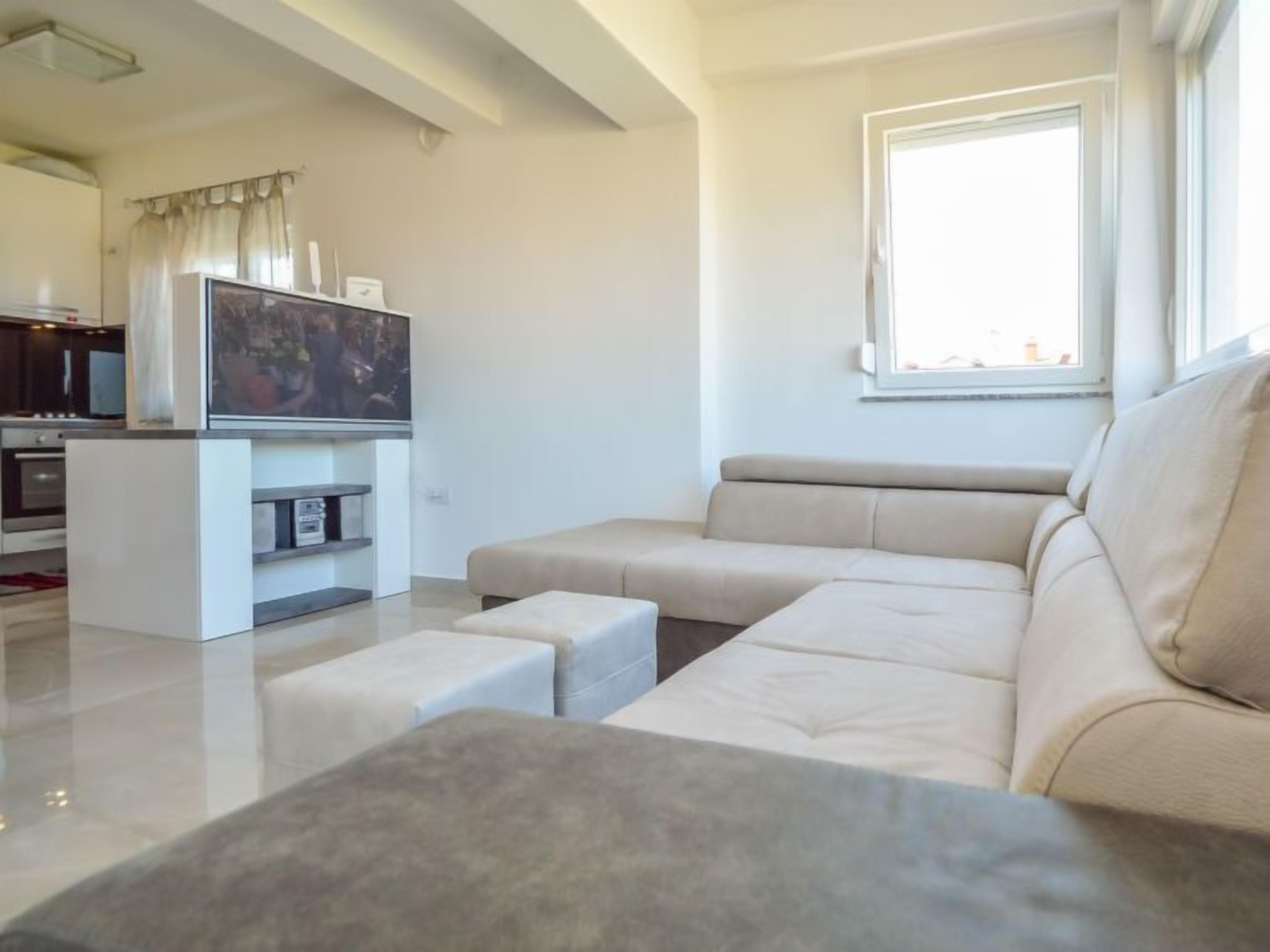 A modern living room with a beige sectional sofa, a flat-screen TV on a white entertainment unit, and a large window providing natural light.