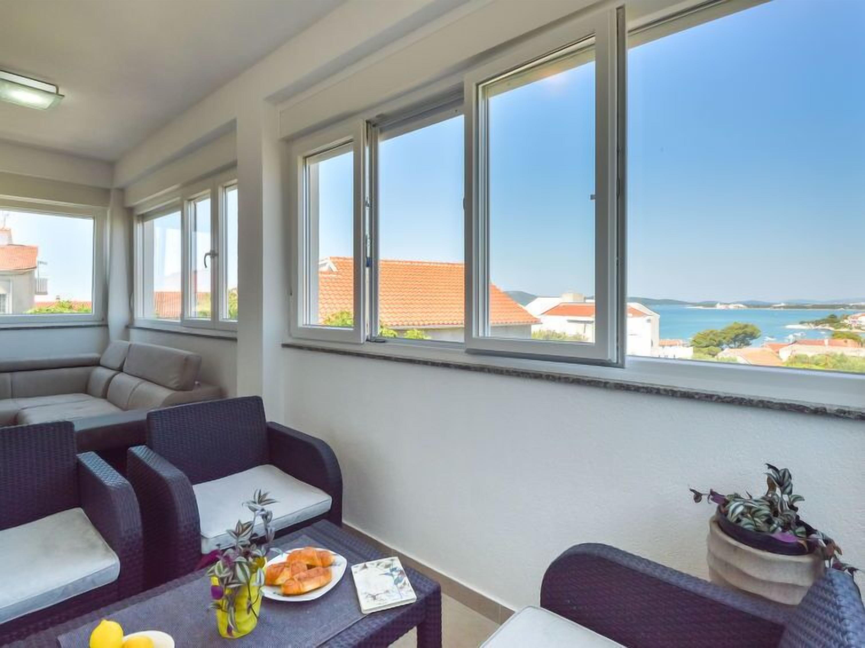 A balcony with a view of the sea, wicker furniture, and a table with a plate of food and a vase of flowers.
