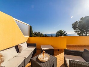 A balcony with a yellow wall, wicker furniture, and a clear view of the sea and trees. - Cadet Rousse by Interhome (Le Lavandou)