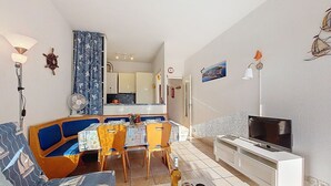 Ein Wohnzimmer mit einer Couch, einem Esstisch, Stühlen, einem Fernseher und einer Küche im Hintergrund.