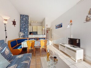Un salon avec un canapé, une table à manger, des chaises, un téléviseur et, au fond, une cuisine.