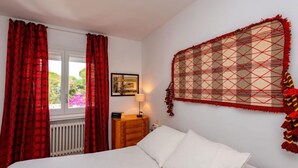 A bedroom with a bed, a nightstand, a window with red curtains, and a wall hanging.