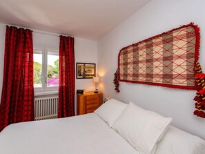 Een slaapkamer met een bed, nachtkastje, raam met rode gordijnen en een wanddecoratie.