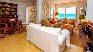 A living room with a white sofa, a wooden dining table, and chairs. There is a view of the ocean through large windows.
