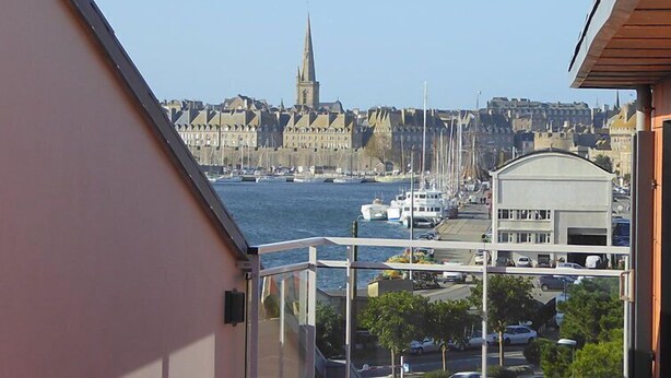 Vista da un balcone su un porto con barche, una guglia di chiesa e una fila di edifici.