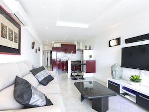 A modern living room with a sofa, coffee table, and TV. There is a kitchen in the background with red cabinets and a dining area.